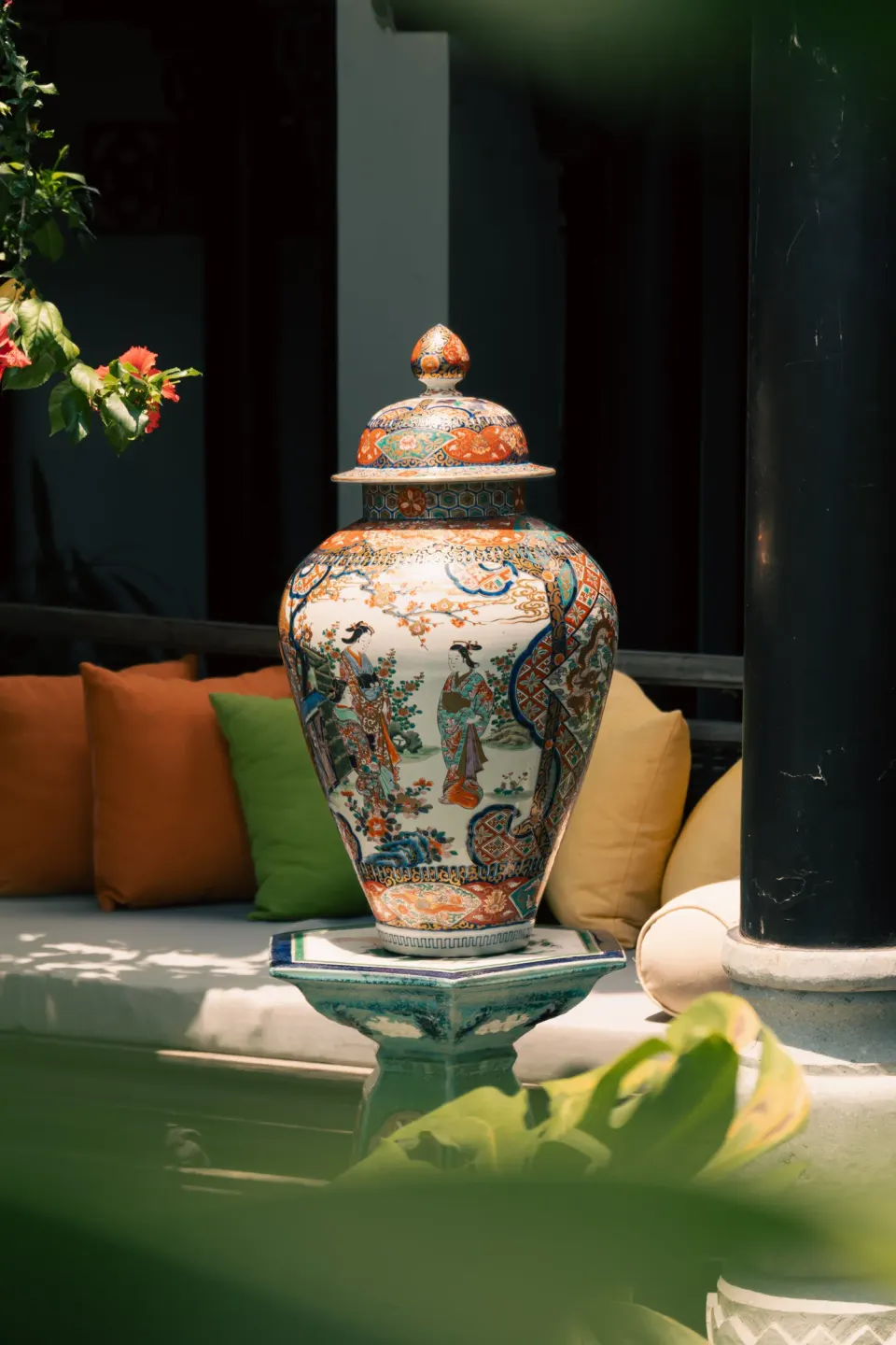 A painted lidded jar on a daybed among orange and green cushions.
