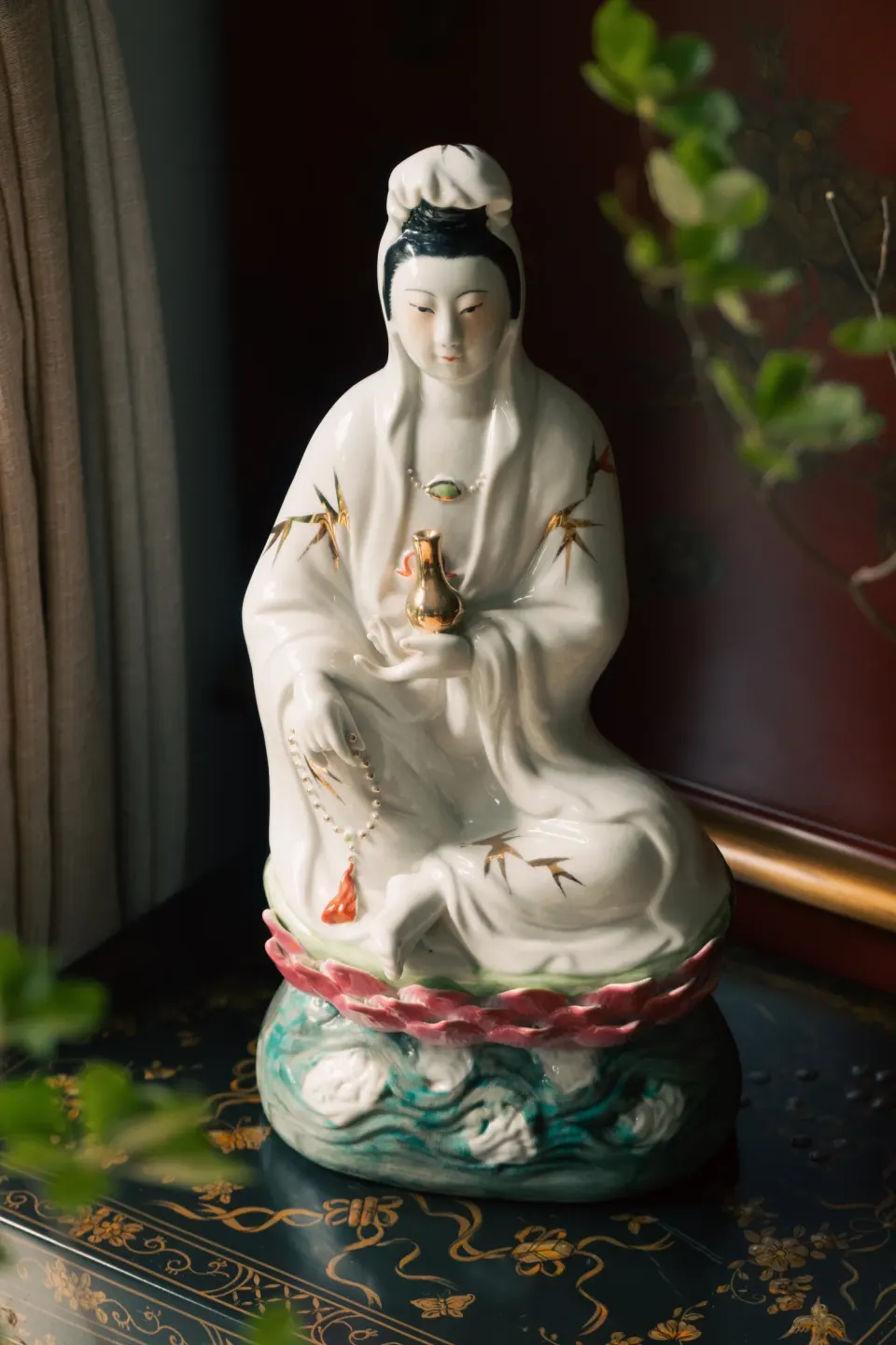 A white porcelain Guanyin seated on a lotus base, on a black lacquer table with gold motifs.