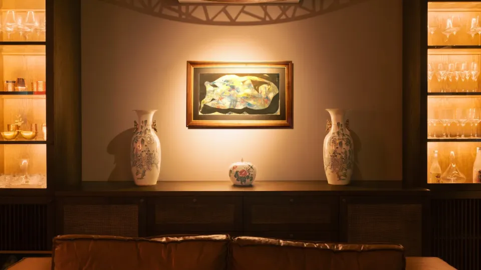 A lamp-lit console under a framed painting, with two porcelain vases, between shelves of glasses.