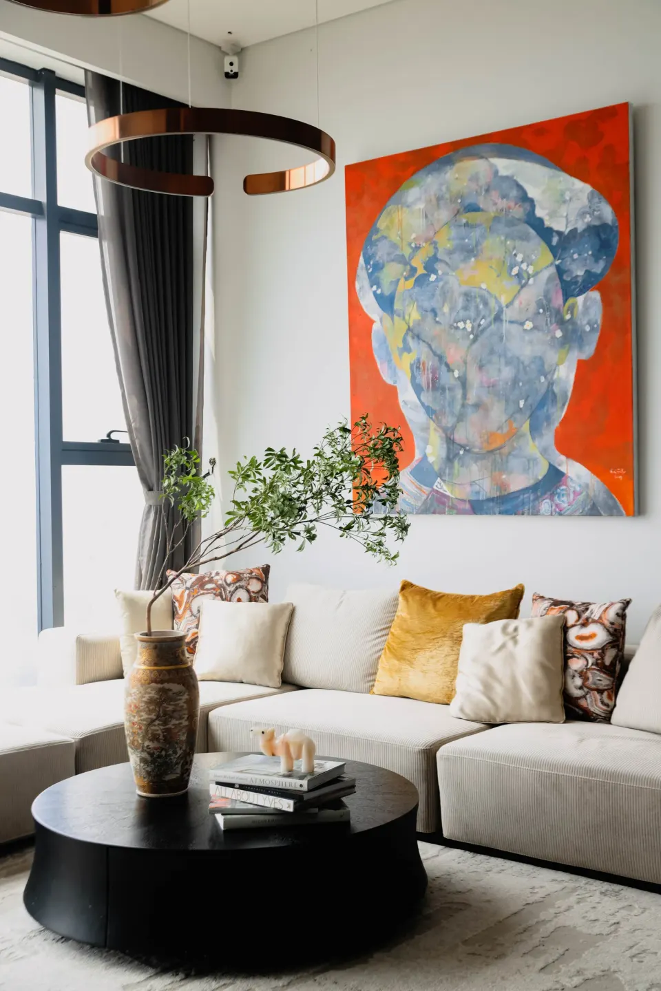 A modern living room: cream sofa, black round table and an orange painting by tall windows.