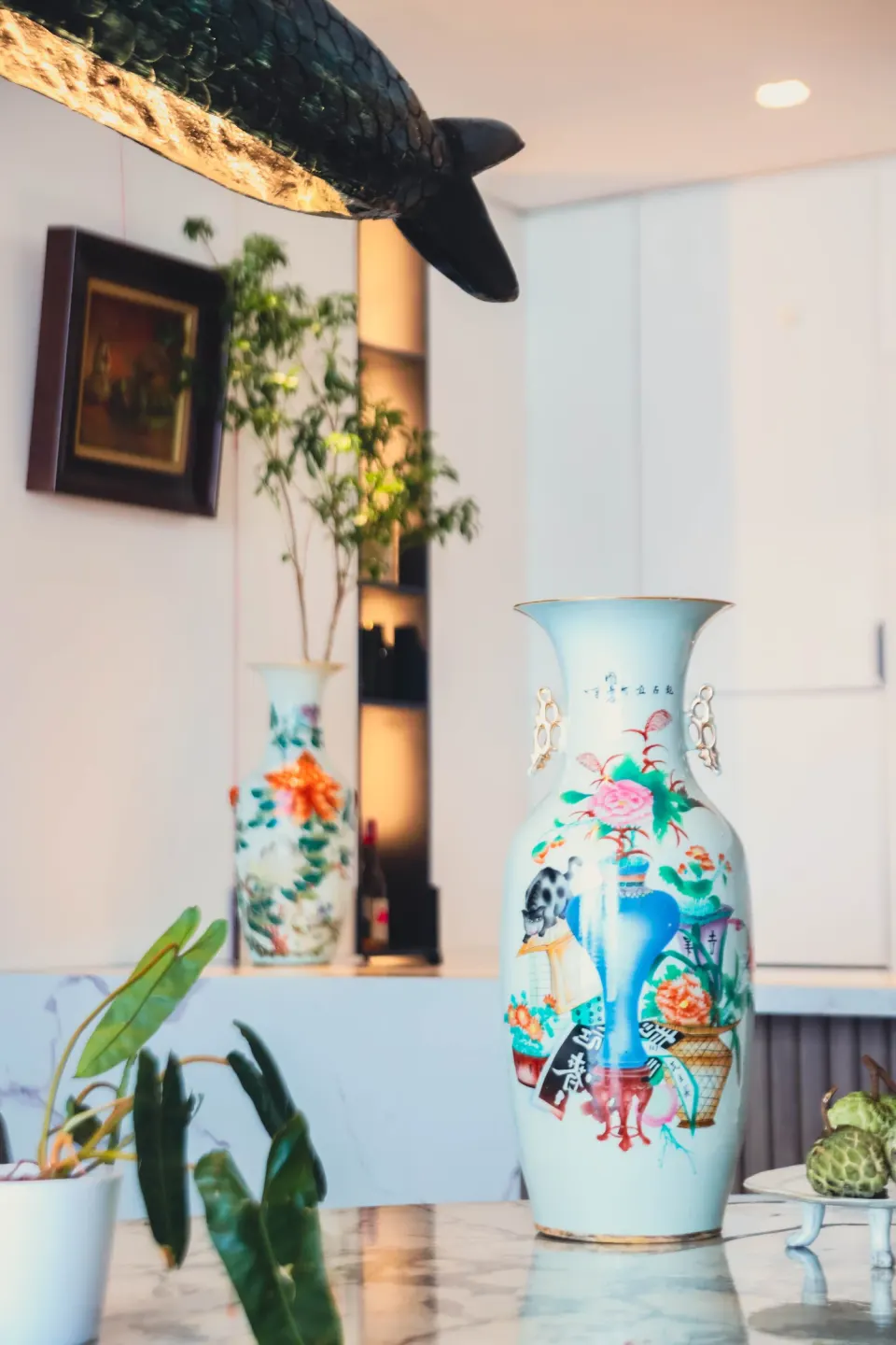 A tall painted vase in front of dark shelves, a sculpture of a fish hanging above.