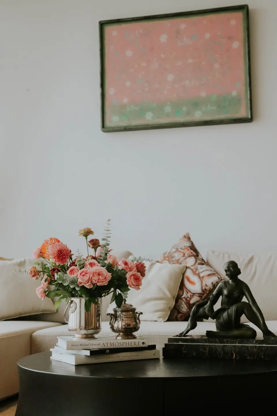 A pink painting above a bouquet and a bronze figure on a coffee table.