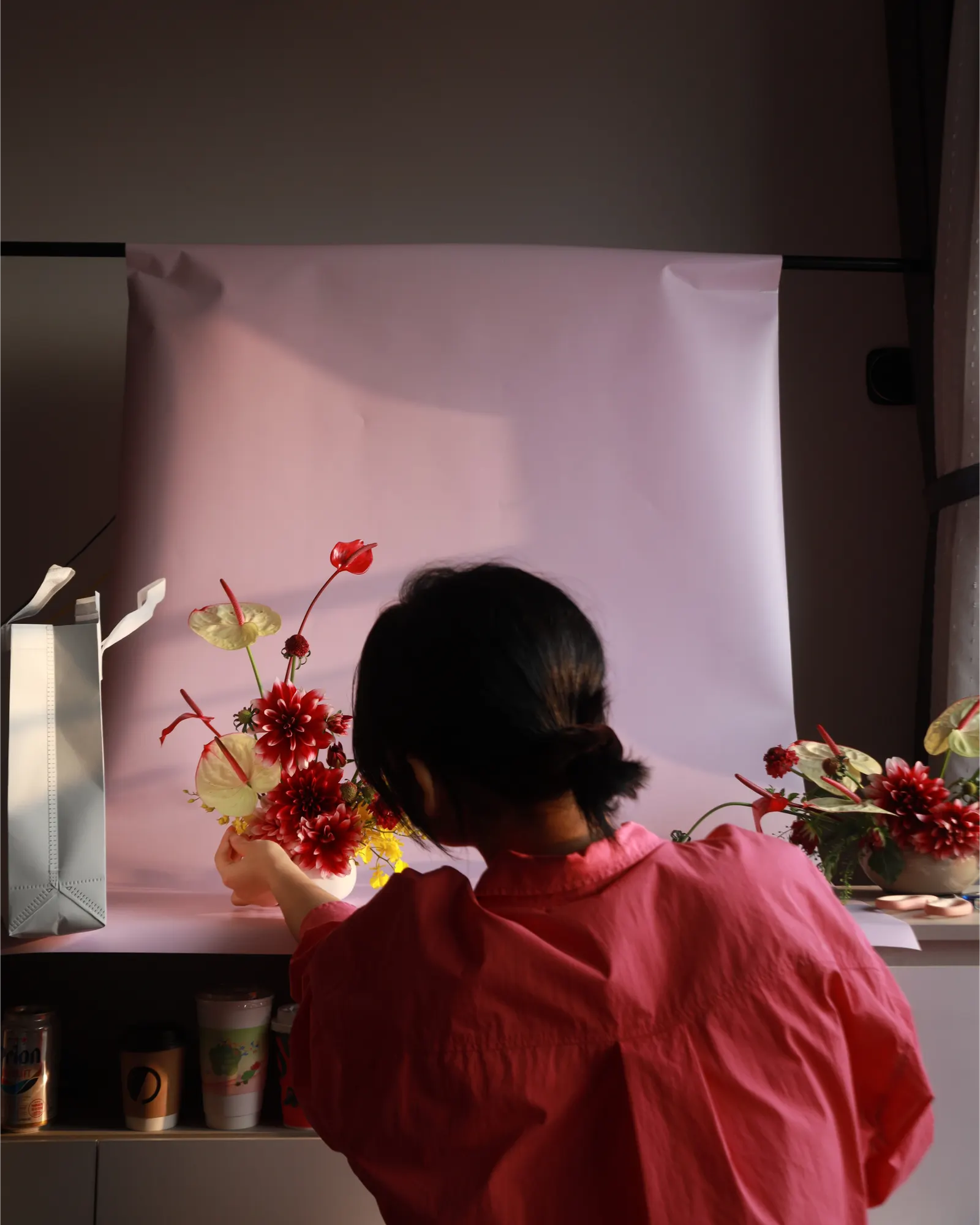 A woman in a pink shirt, seen from behind, arranging flowers in front of a pale pink paper backdrop.
