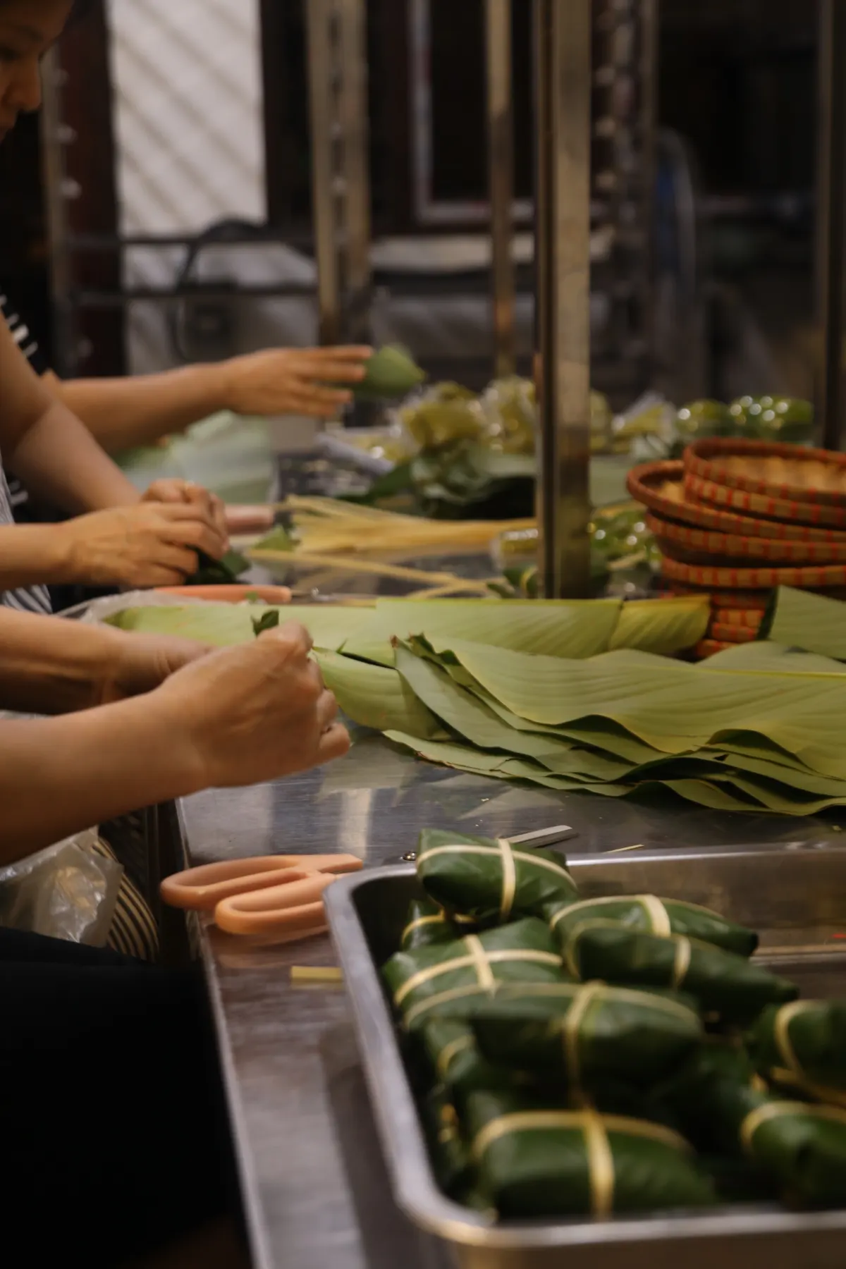 Hands folding green leaves around cakes at a steel counter, with wrapped parcels in a tray in front.
