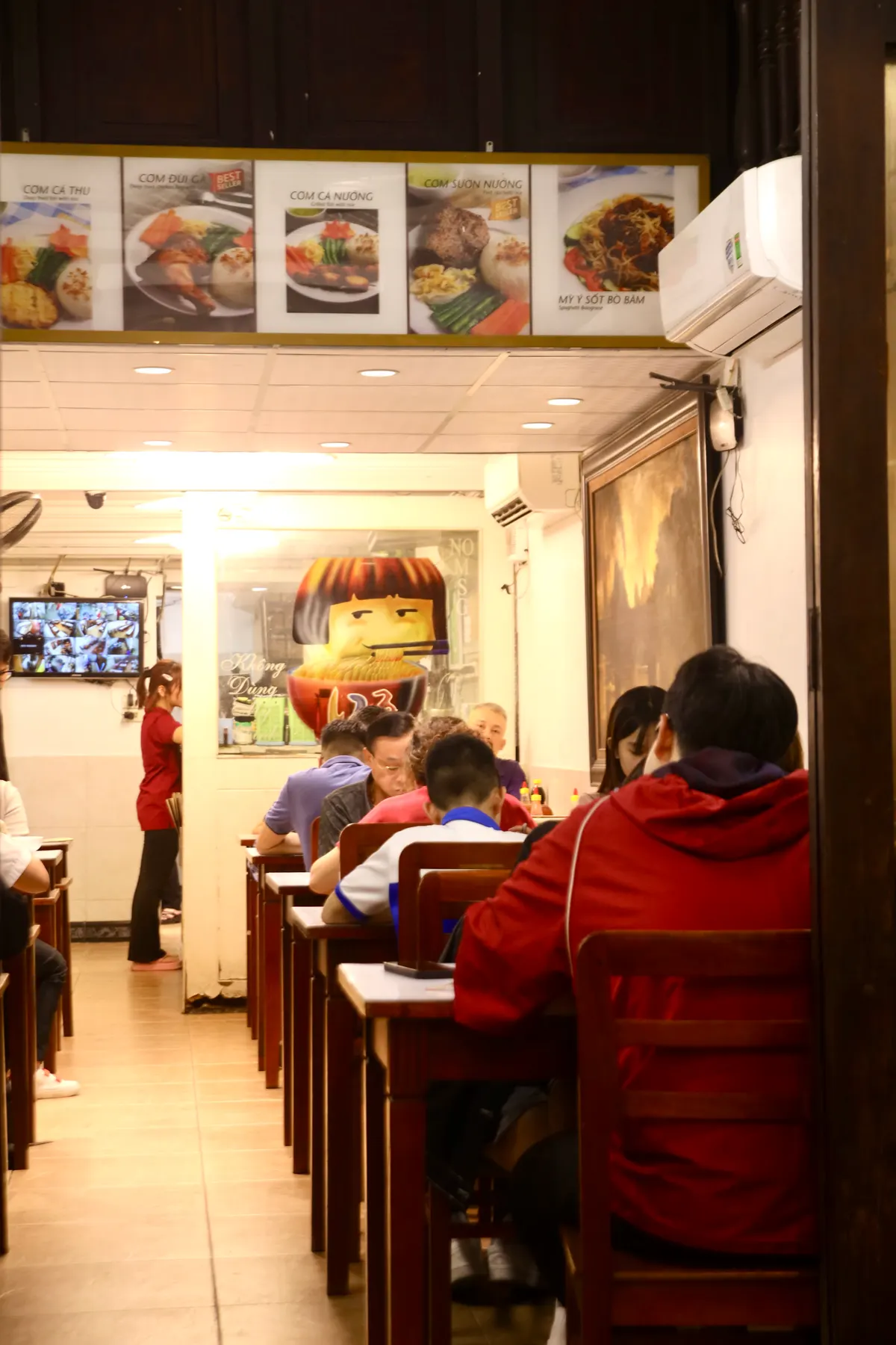 A narrow, bright restaurant with customers at wooden tables, a waitress in red and a cartoon face painted on the wall.