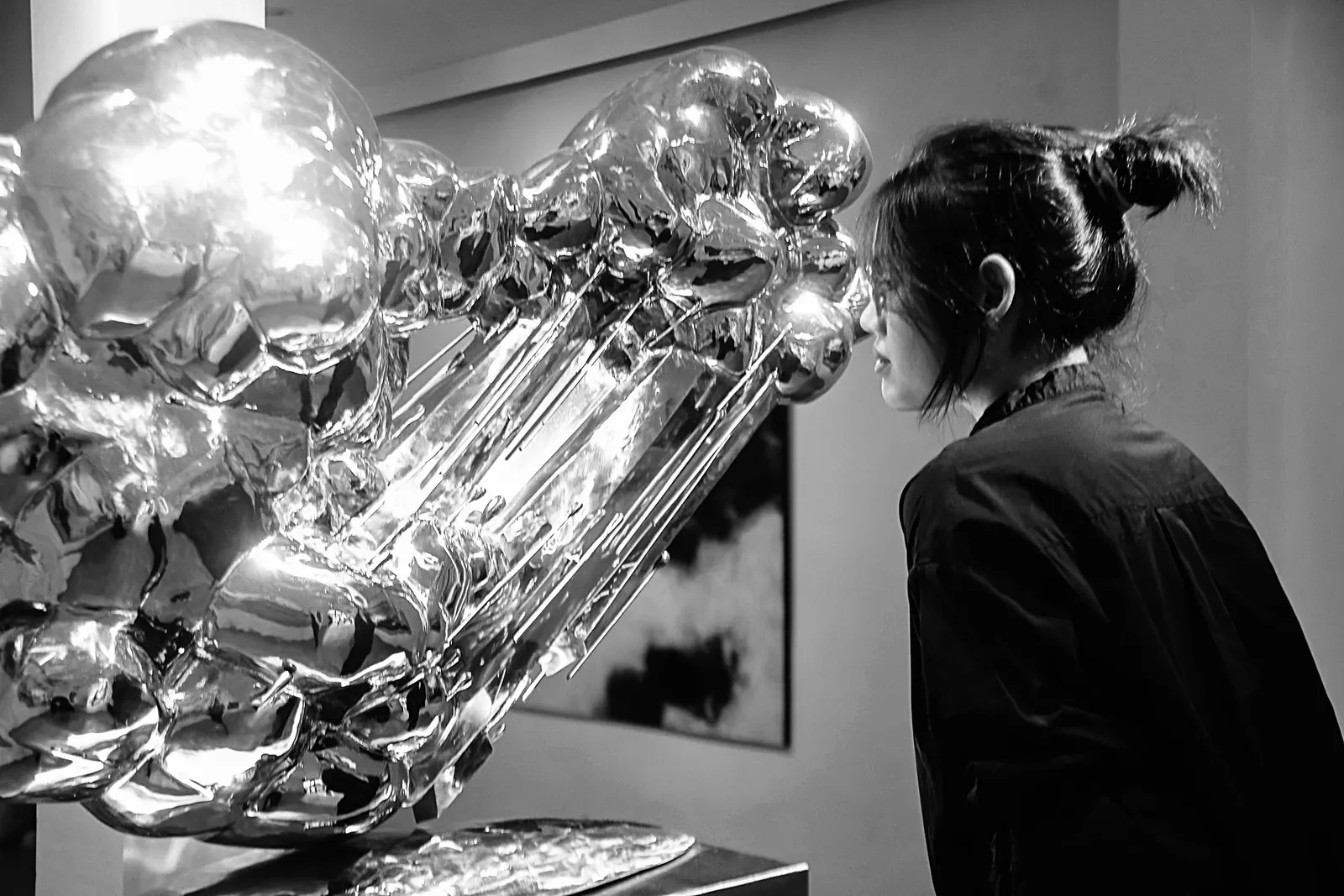 Cam Anh in profile, hair in a clip, looking closely at a mirror-polished silver sculpture in a pale gallery.