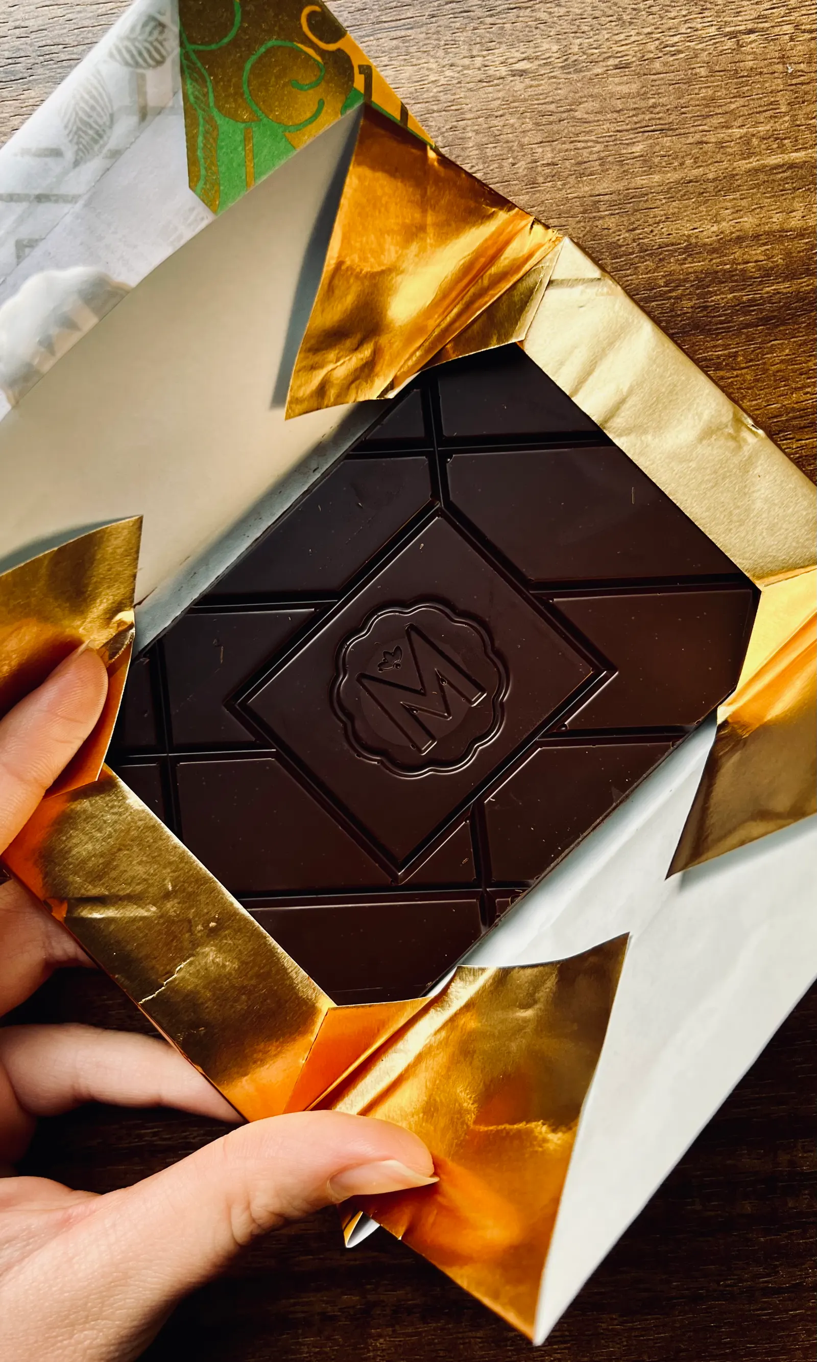 A hand opening gold foil to reveal a dark chocolate bar stamped with an M, on a wooden table.