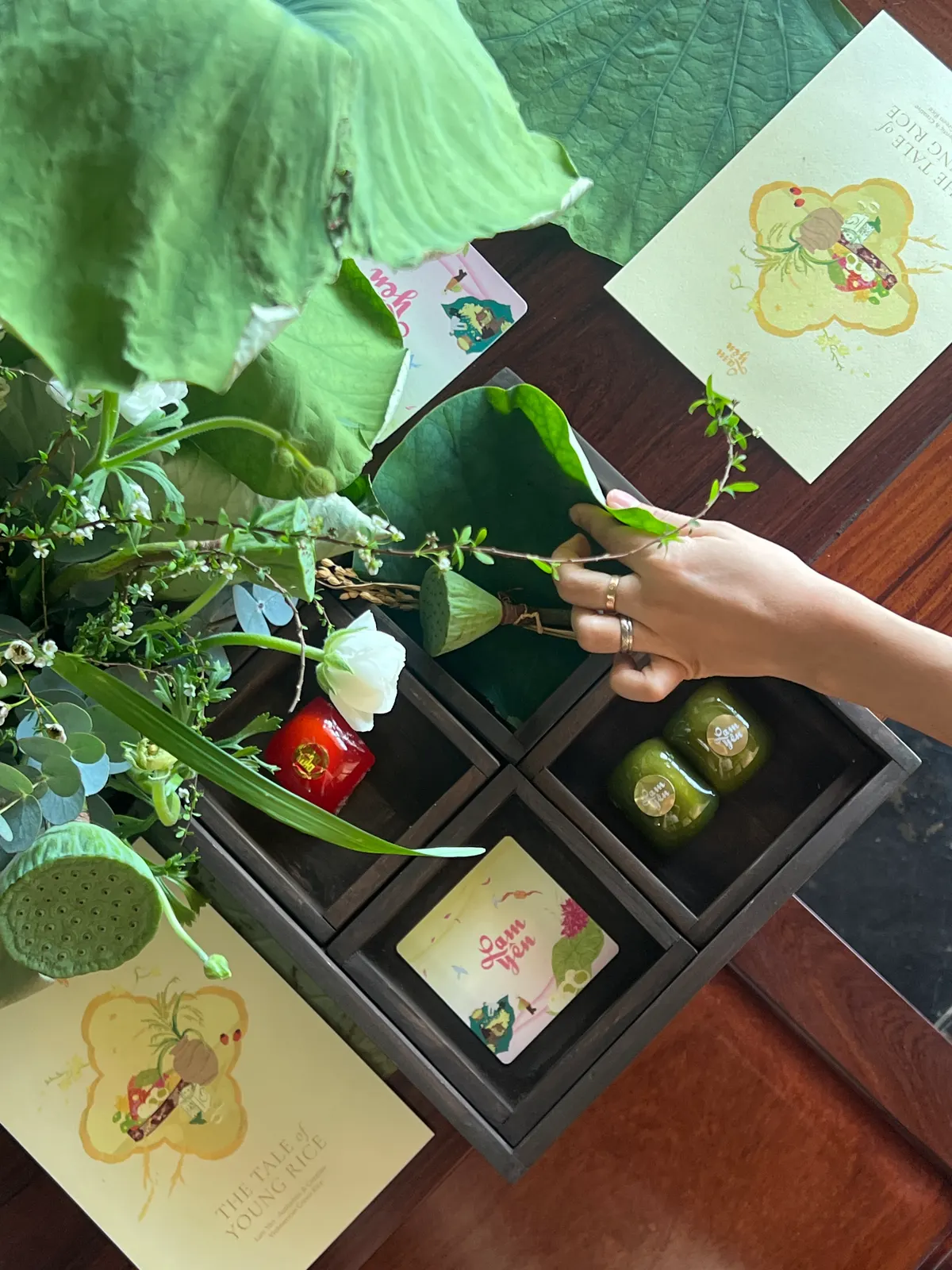 A hand arranging a lotus leaf and flowers over a dark wooden tray of small cakes, with printed cards beside it.
