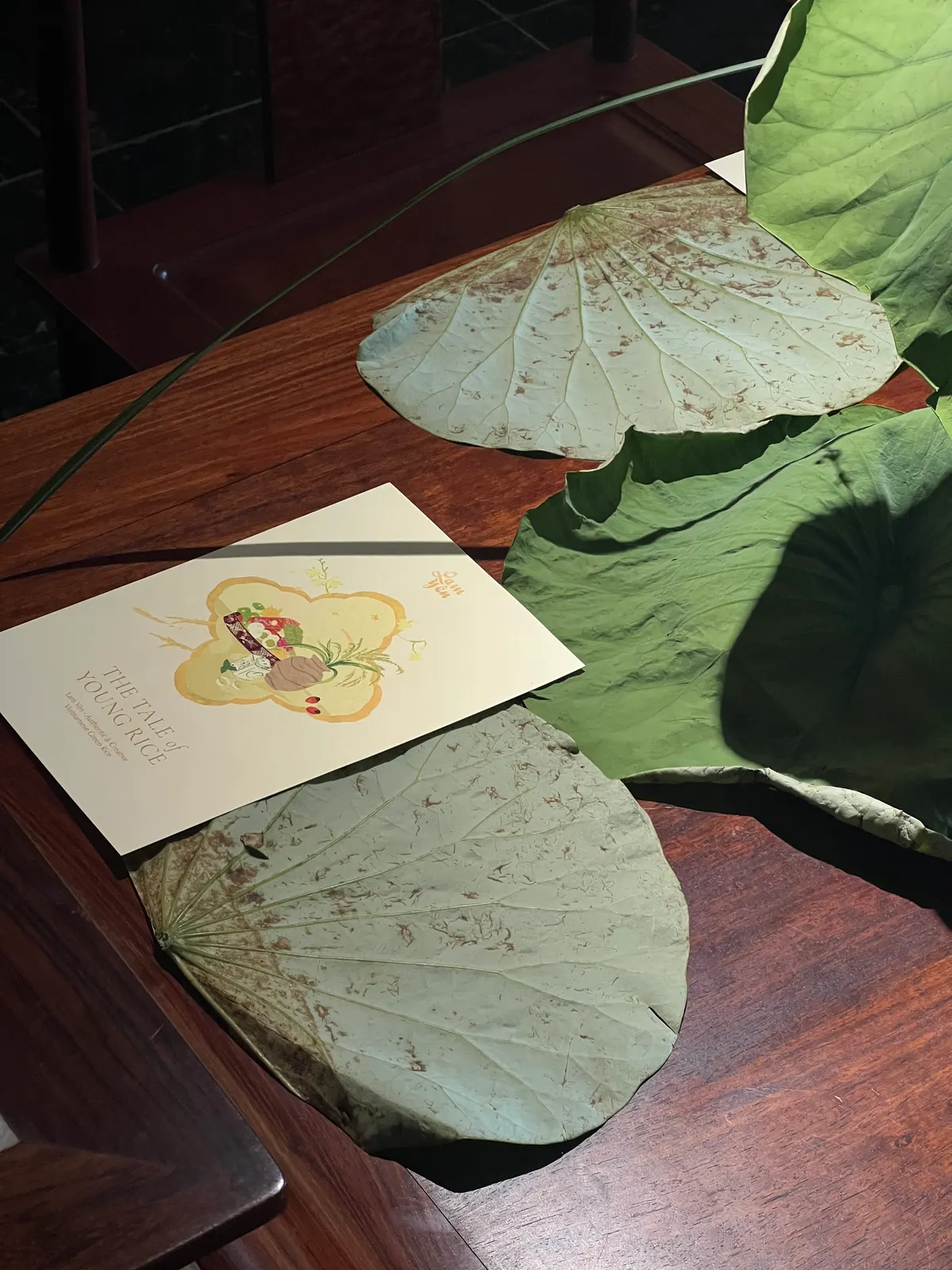 A printed invitation card lying on a dark wooden table between dried lotus leaves.