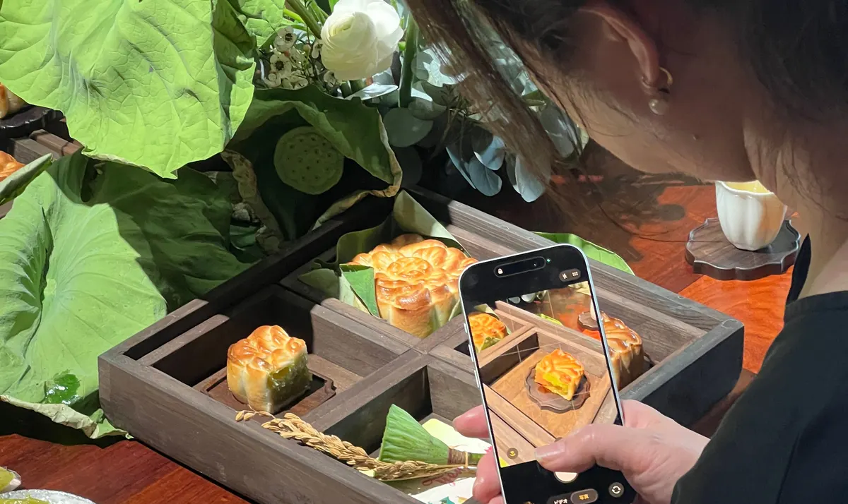 A guest photographing a wooden tray of mooncakes with her phone, among lotus leaves and flowers.