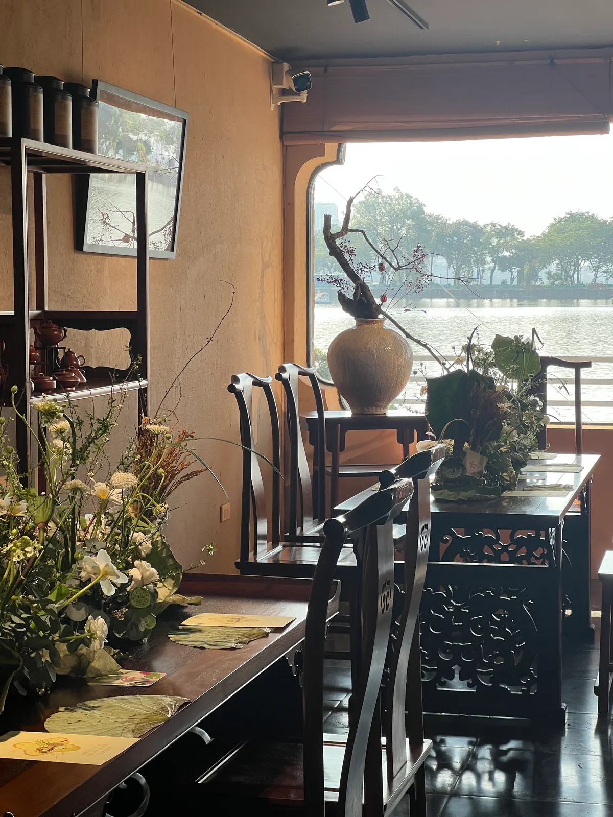 A quiet dining room with carved wooden chairs and a long table of flowers, beside a window over a lake.