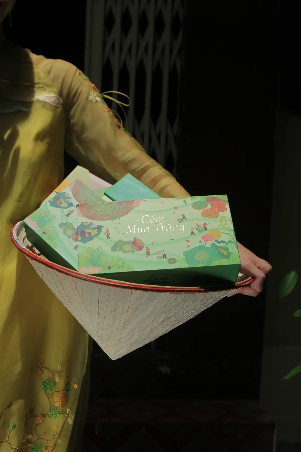 A conical hat used as a tray, holding a green Cốm Mùa Trăng box, with a woman in a yellow áo dài behind it.