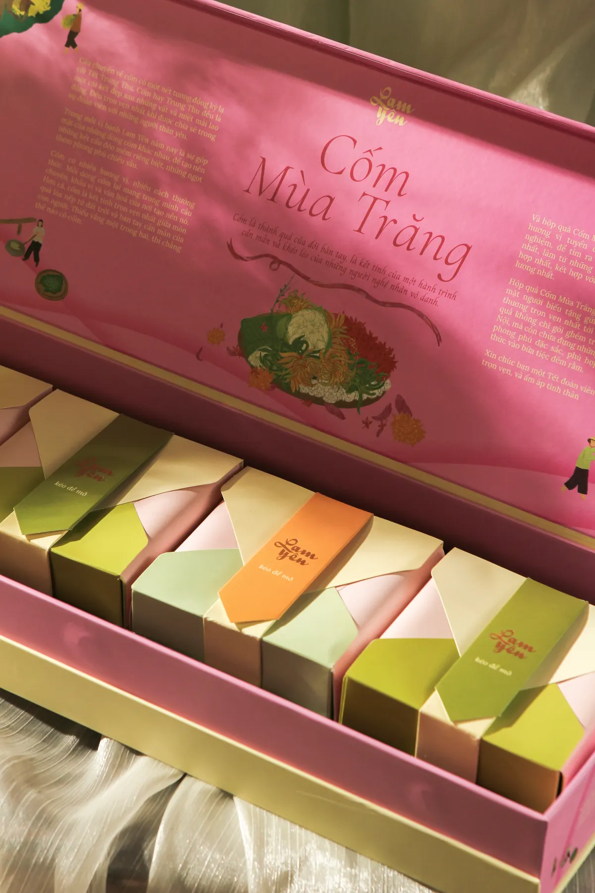An open pink box of pastel mooncake boxes under a printed lid lettered “Cốm Mùa Trăng”.