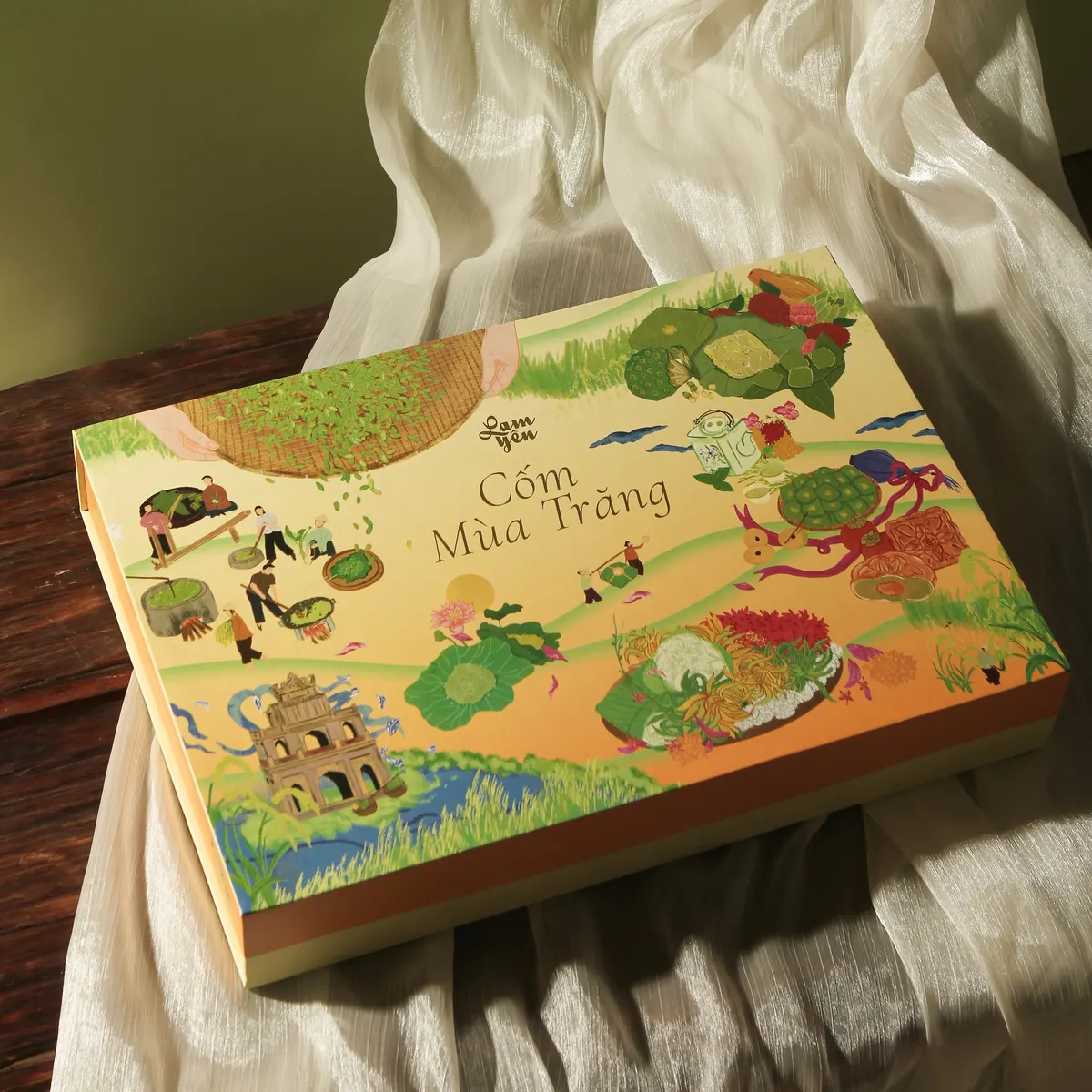 The Cốm Mùa Trăng box, covered in illustrated village scenes, lying on a cream cloth on a wooden table.