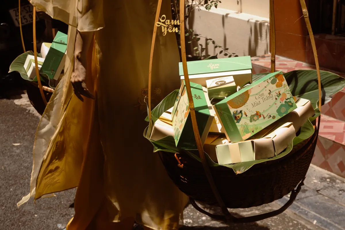 A shoulder-pole basket heaped with green and pink gift boxes, carried by a woman in yellow.