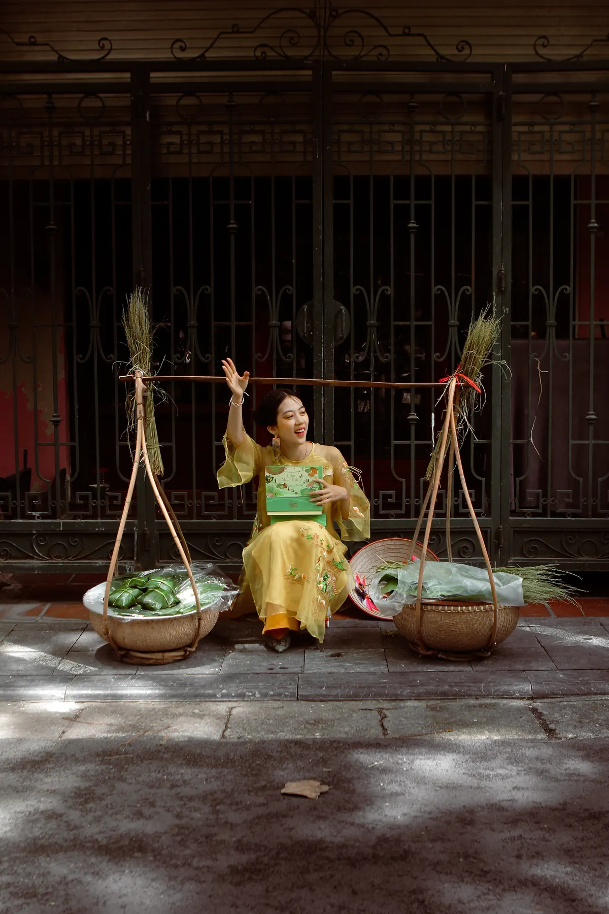 A woman in yellow sitting on the pavement between two baskets of leaves, one arm raised.