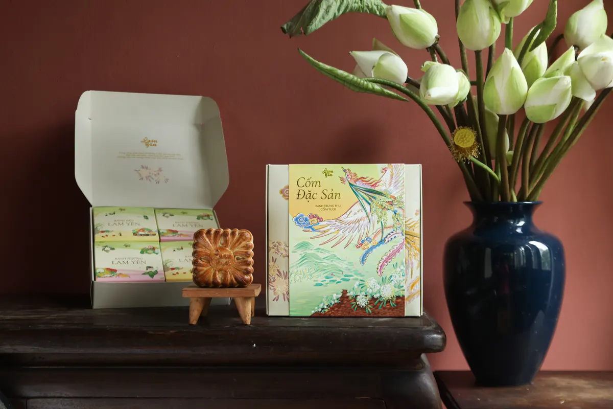 An open box of Cốm Đặc Sản beside a closed one and a mooncake on a small stand, next to a vase of white lotus buds, against a dusty-pink wall.