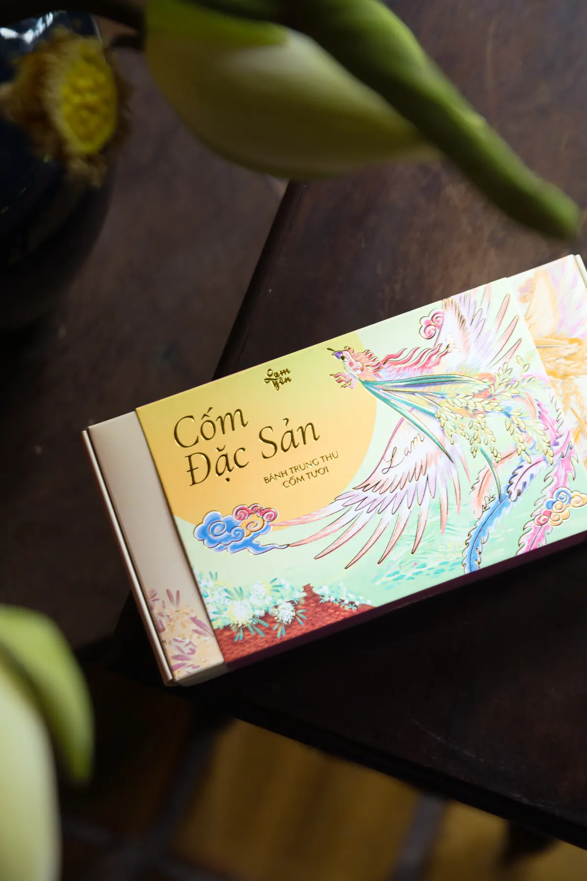 The Cốm Đặc Sản box lying at an angle on a dark wooden table, white lotus buds above it.