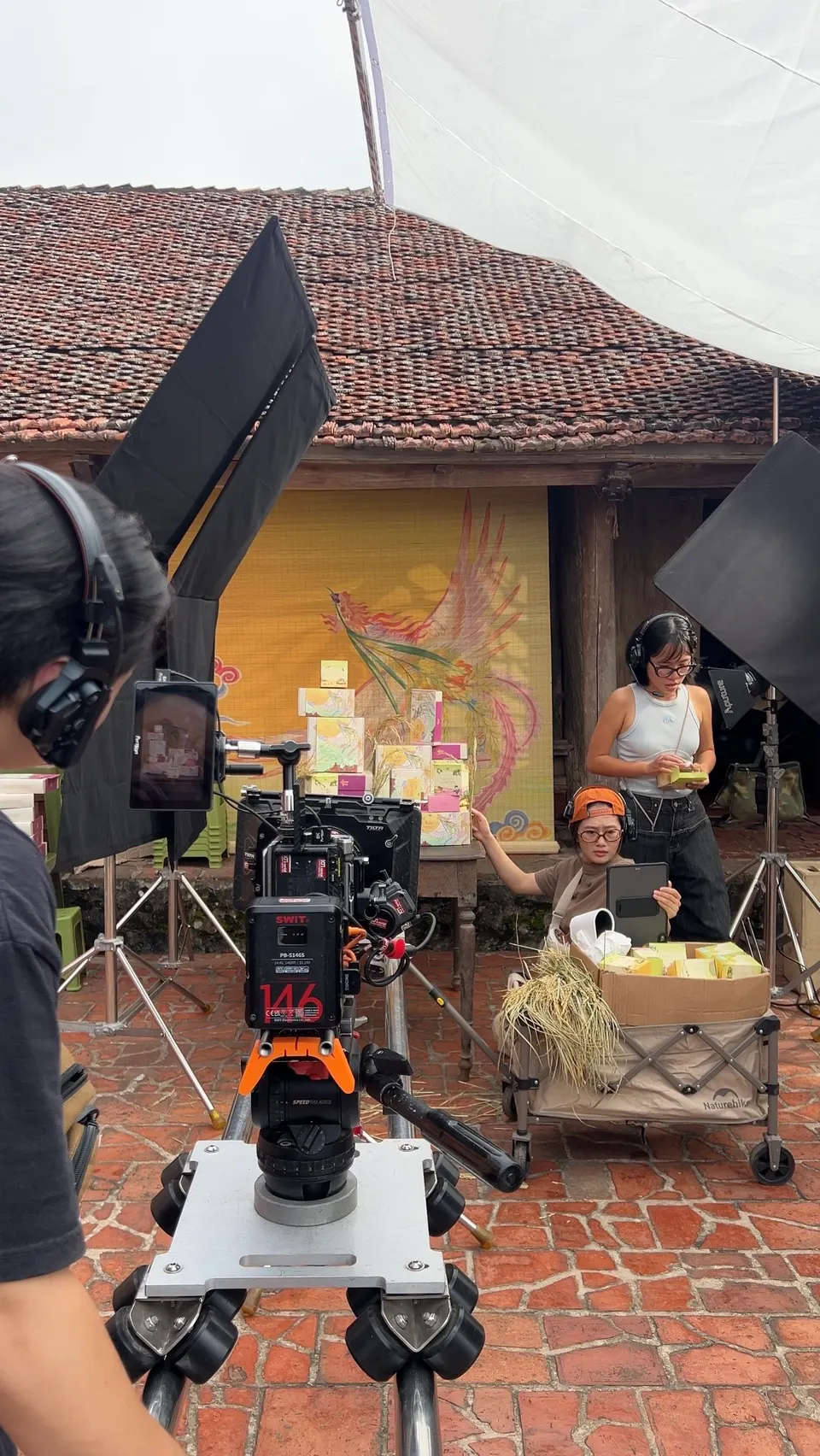 A film camera on a rig in a courtyard, facing a table of gift boxes and a woman sitting behind it.