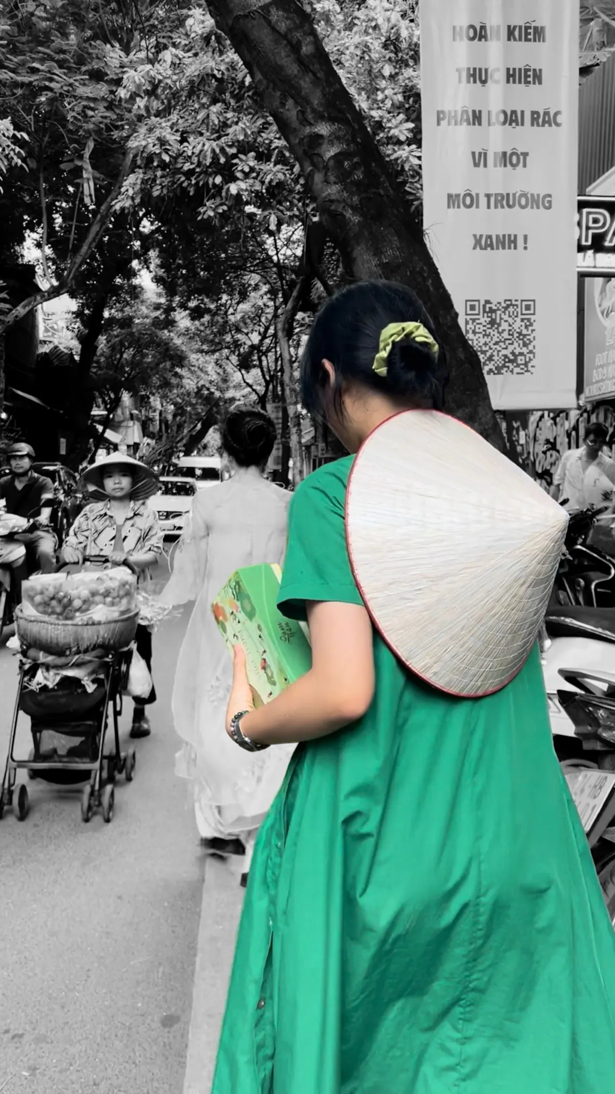 A woman in a green dress and a conical hat, seen from behind on a busy Hanoi street, carrying a green gift box; the street is in black and white.