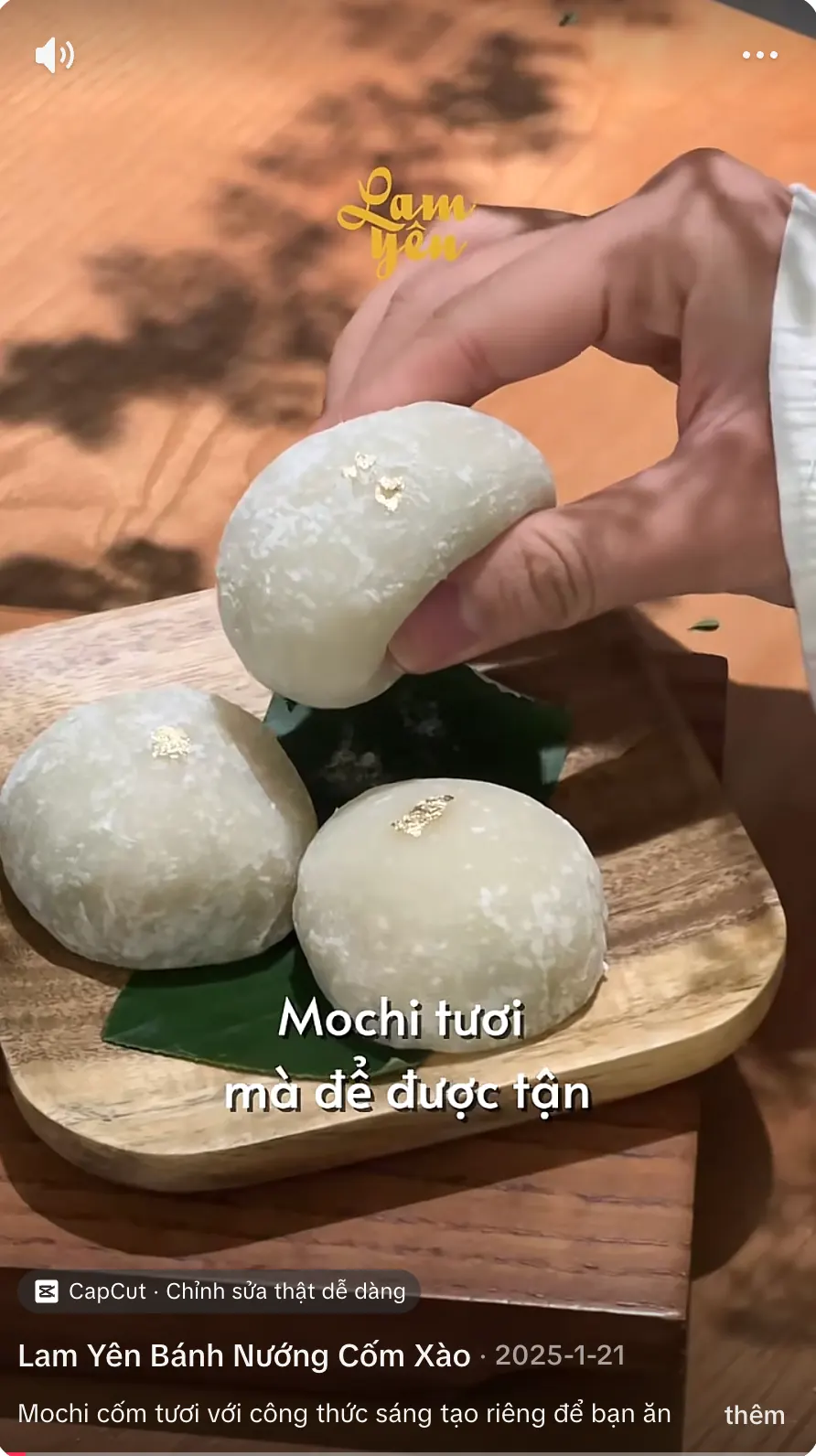 A video still: a hand holding a fresh white mochi dusted with gold flakes above two more on a wooden board with a banana leaf, with the words “Mochi tươi mà dễ được tận”.
