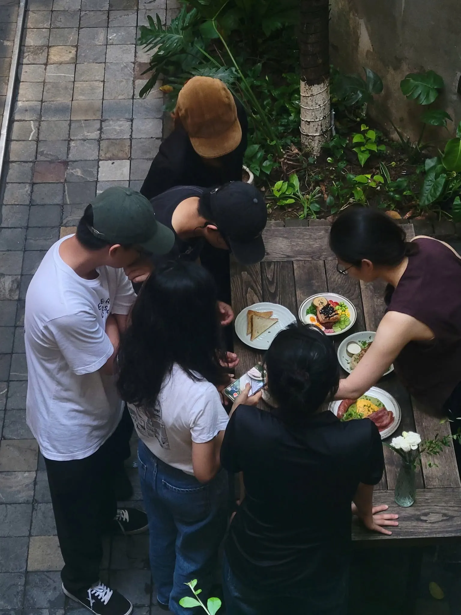 Seen from above, six team members in a café courtyard lean over a wooden table of brunch plates, one of them filming the dishes on a phone.