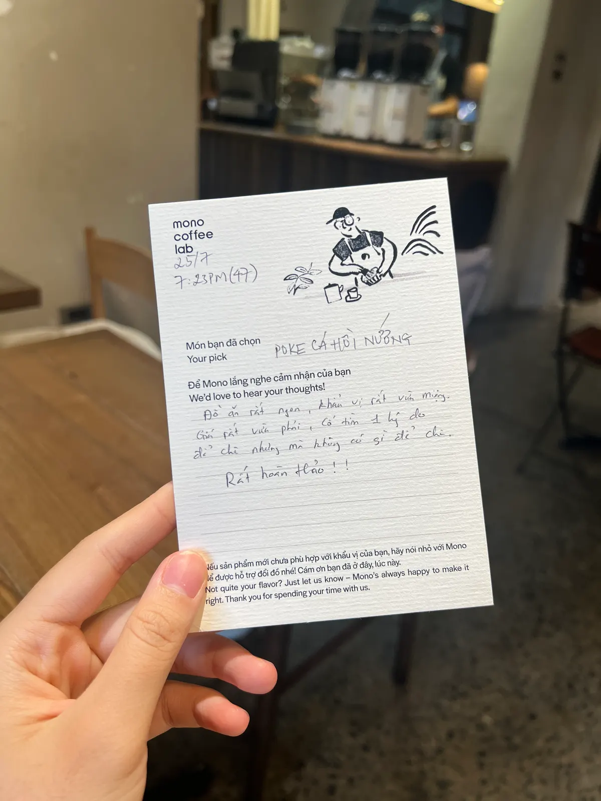 A hand holding up a Mono Coffee Lab guest opinion card, filled in by hand, in the café (card 3 of 9).