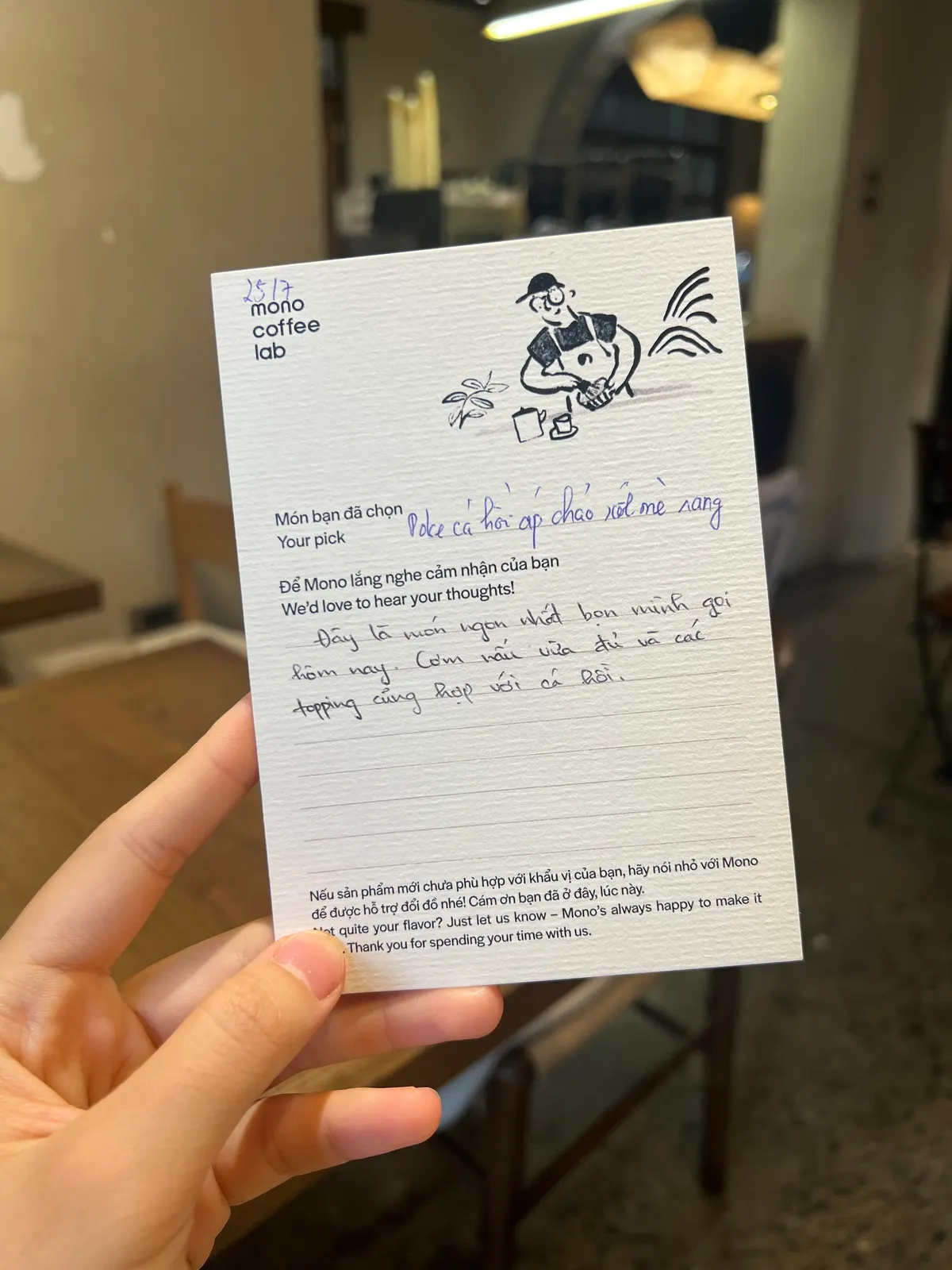 A hand holding up a Mono Coffee Lab guest opinion card, filled in by hand, in the café (card 6 of 9).