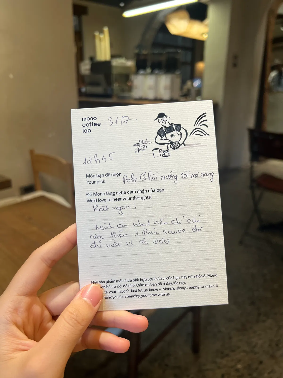 A hand holding up a Mono Coffee Lab guest opinion card, filled in by hand, in the café (card 7 of 9).