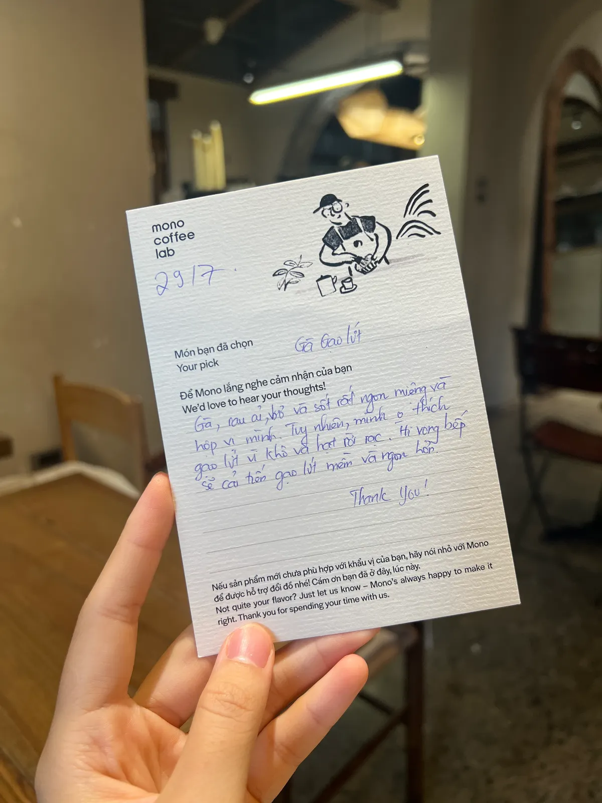A hand holding up a Mono Coffee Lab guest opinion card, filled in by hand, in the café (card 9 of 9).