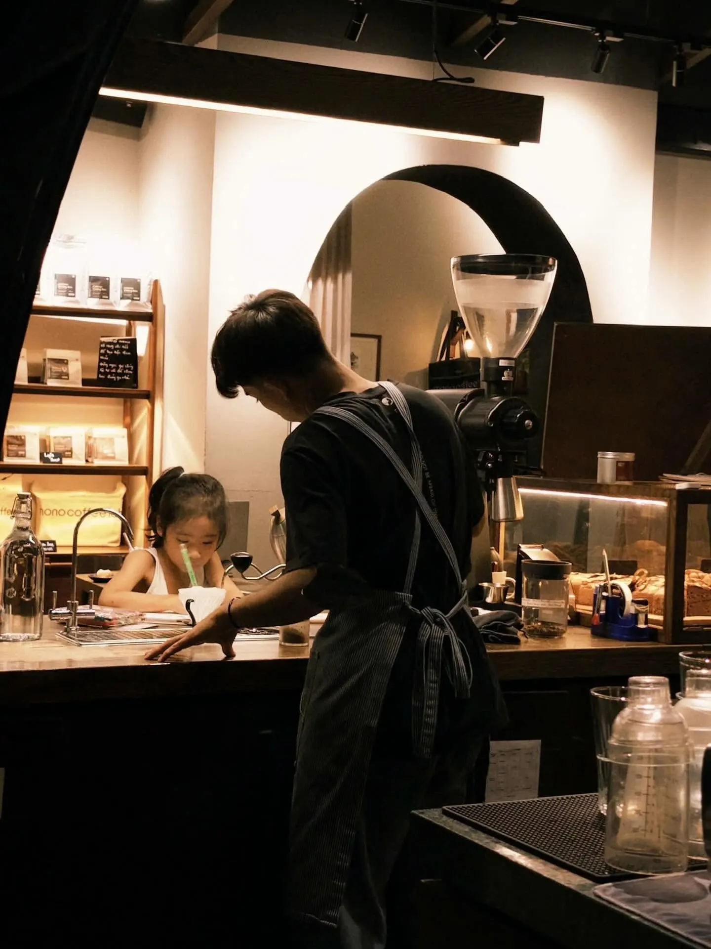 Inside Mono Coffee Lab at night: a barista in a striped apron leans over the counter, a small girl drawing behind it, shelves of coffee bags and a glowing arch in the wall.
