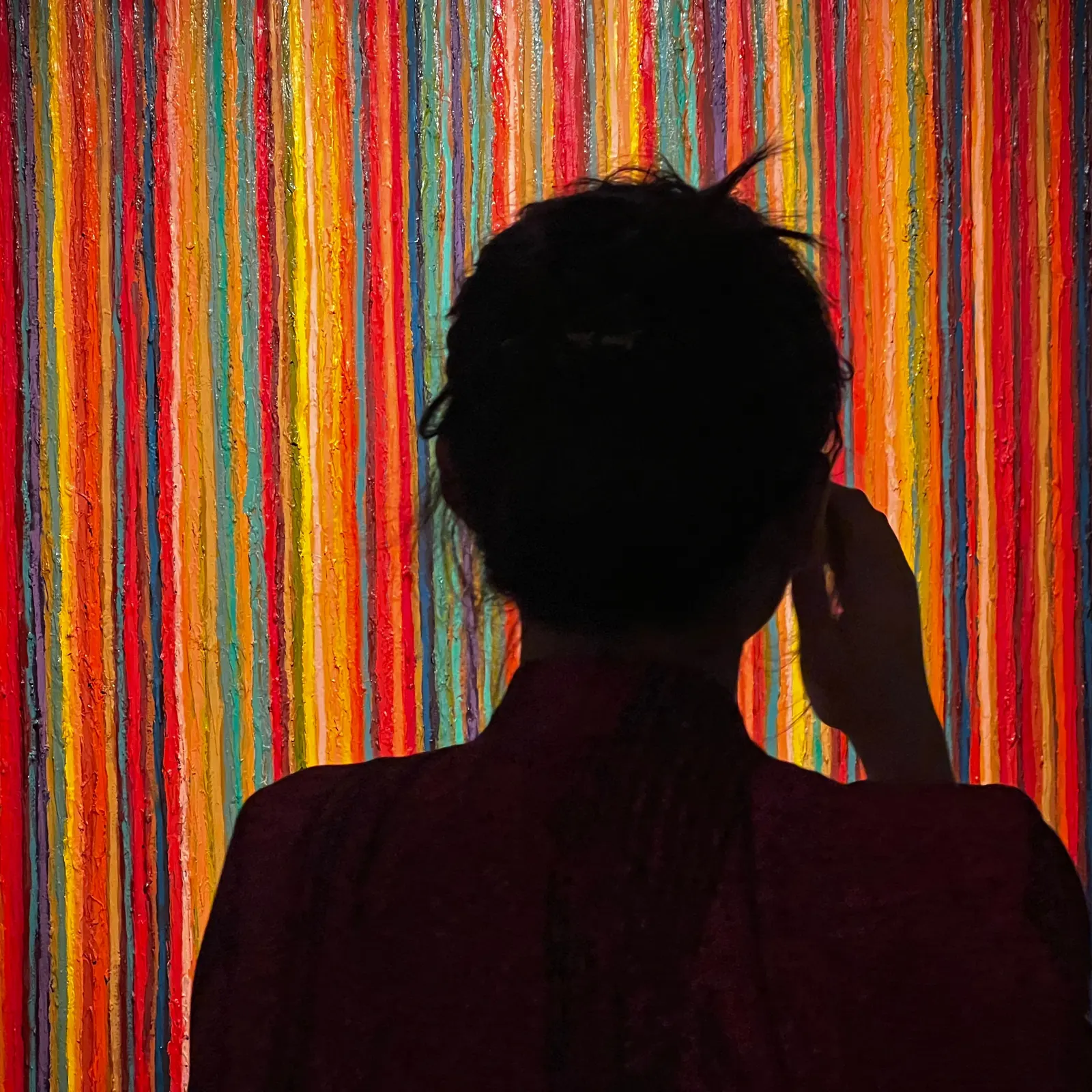 The silhouette of Cam Anh from behind, one hand at her ear, against a wall of thick vertical stripes of colourful paint.