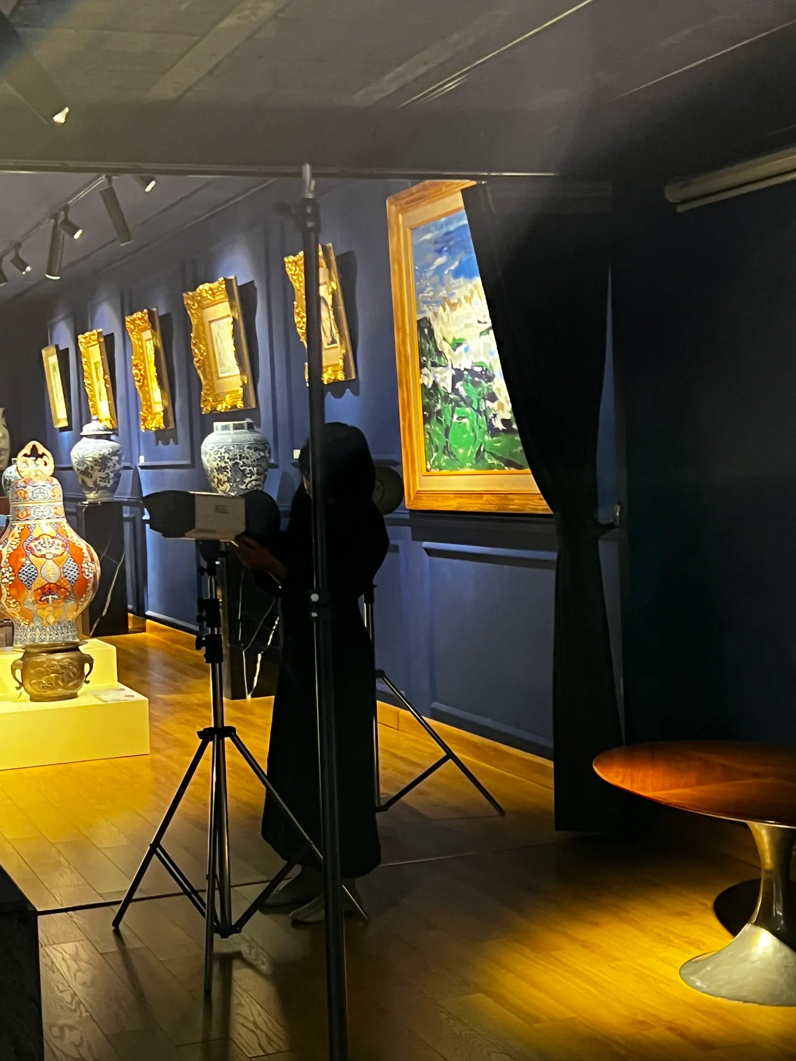 A photographer working in a navy gallery, a single beam of light and a tripod among gilded frames and porcelain.