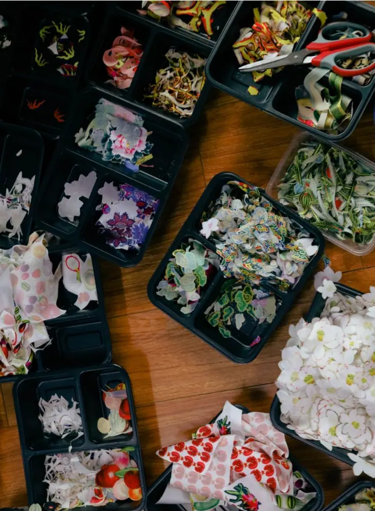 Black trays of cut paper and dried flower petals in many colours on a wooden floor, with scissors.