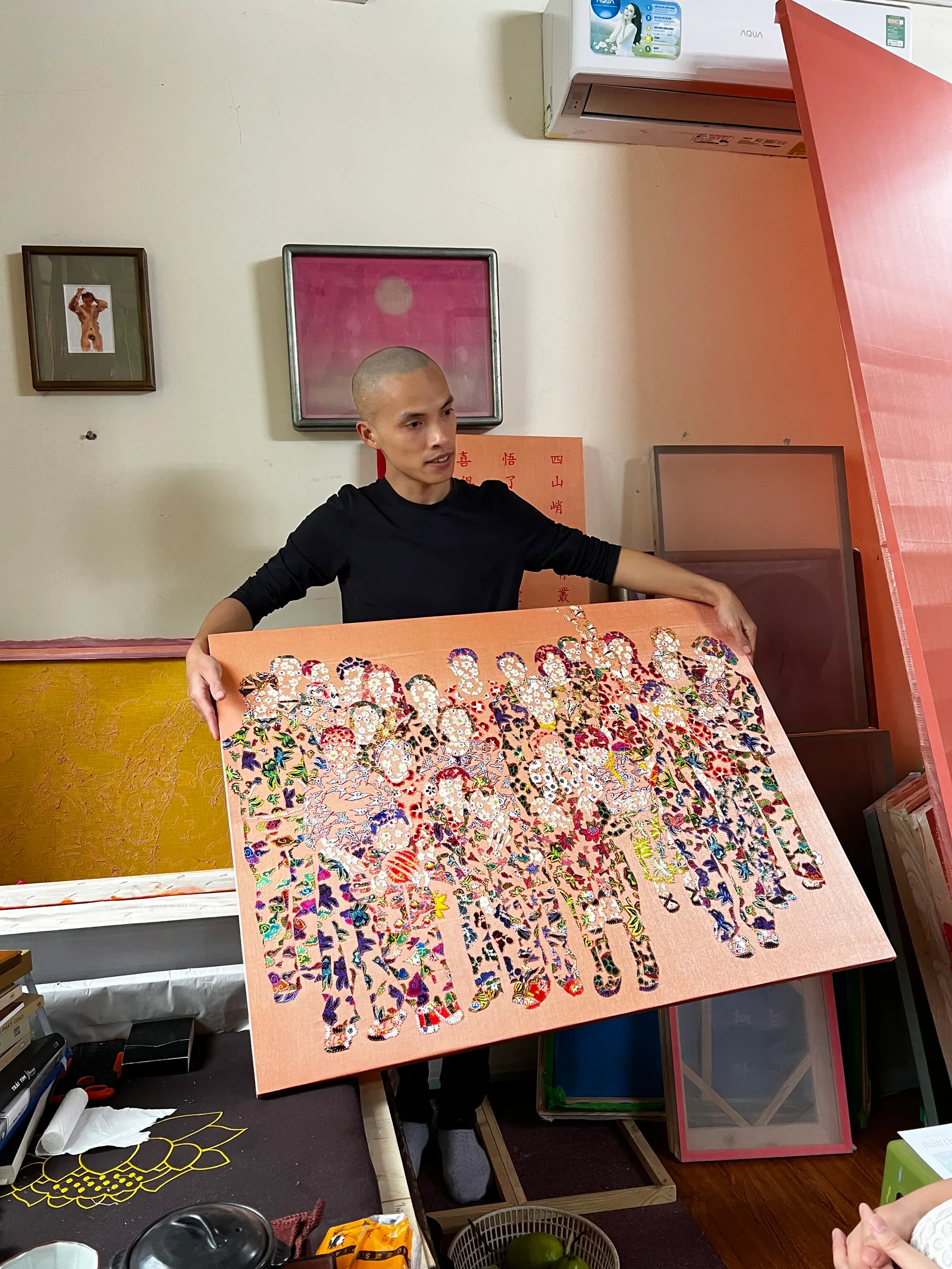 An artist holding up a crowded, colourful painting on an orange panel.