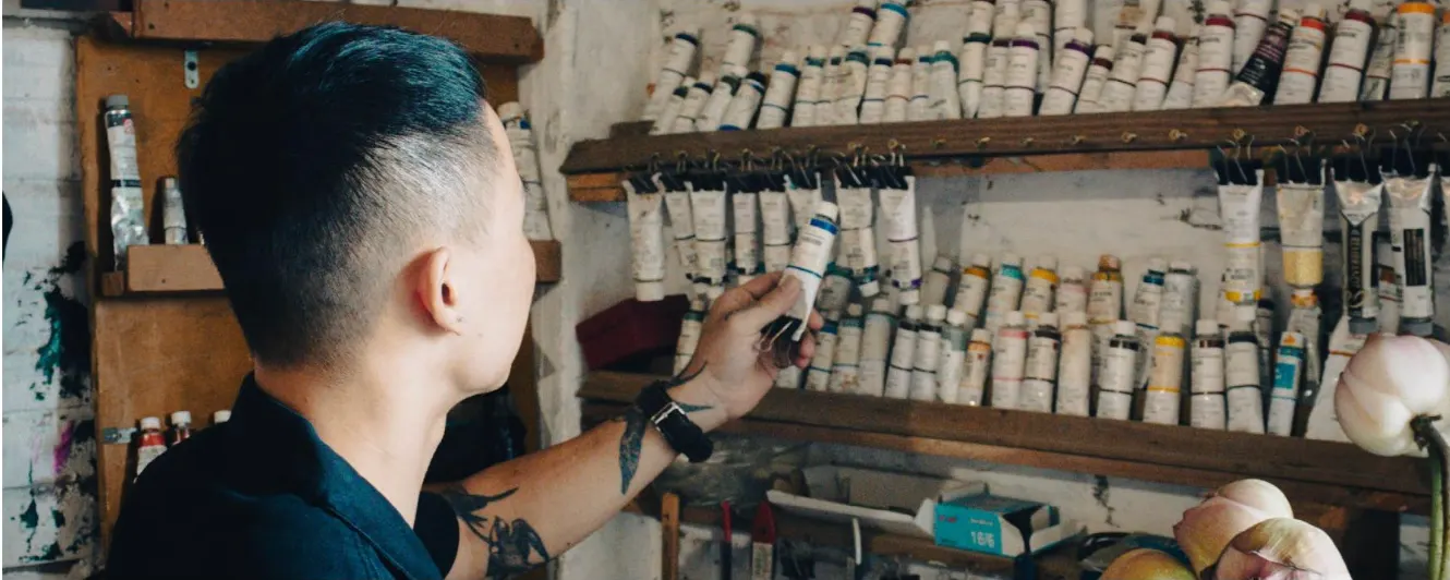 An artist reaching for a tube of paint from a wall of shelves lined with paint tubes.