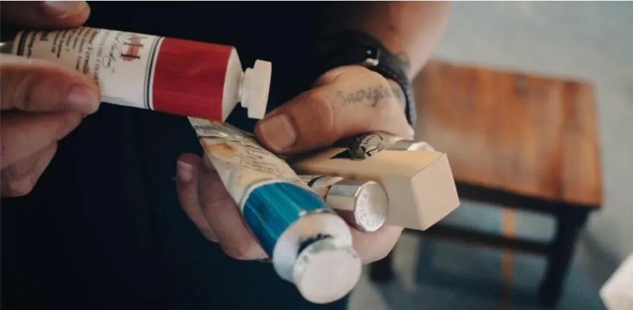 Hands squeezing a tube of red paint and a tube of blue paint at a small wooden table.