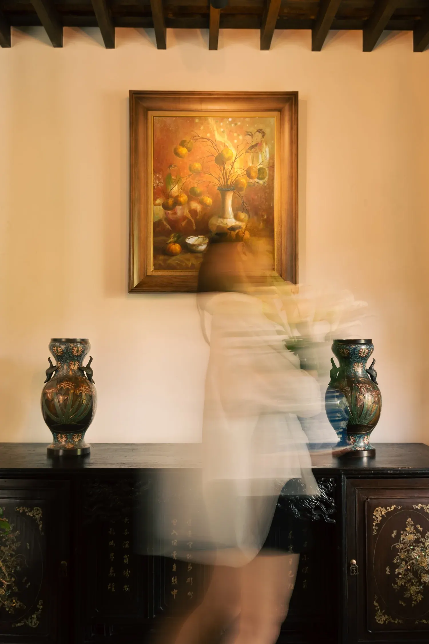 A framed painting above a dark console with two vases, a blurred figure walking past.