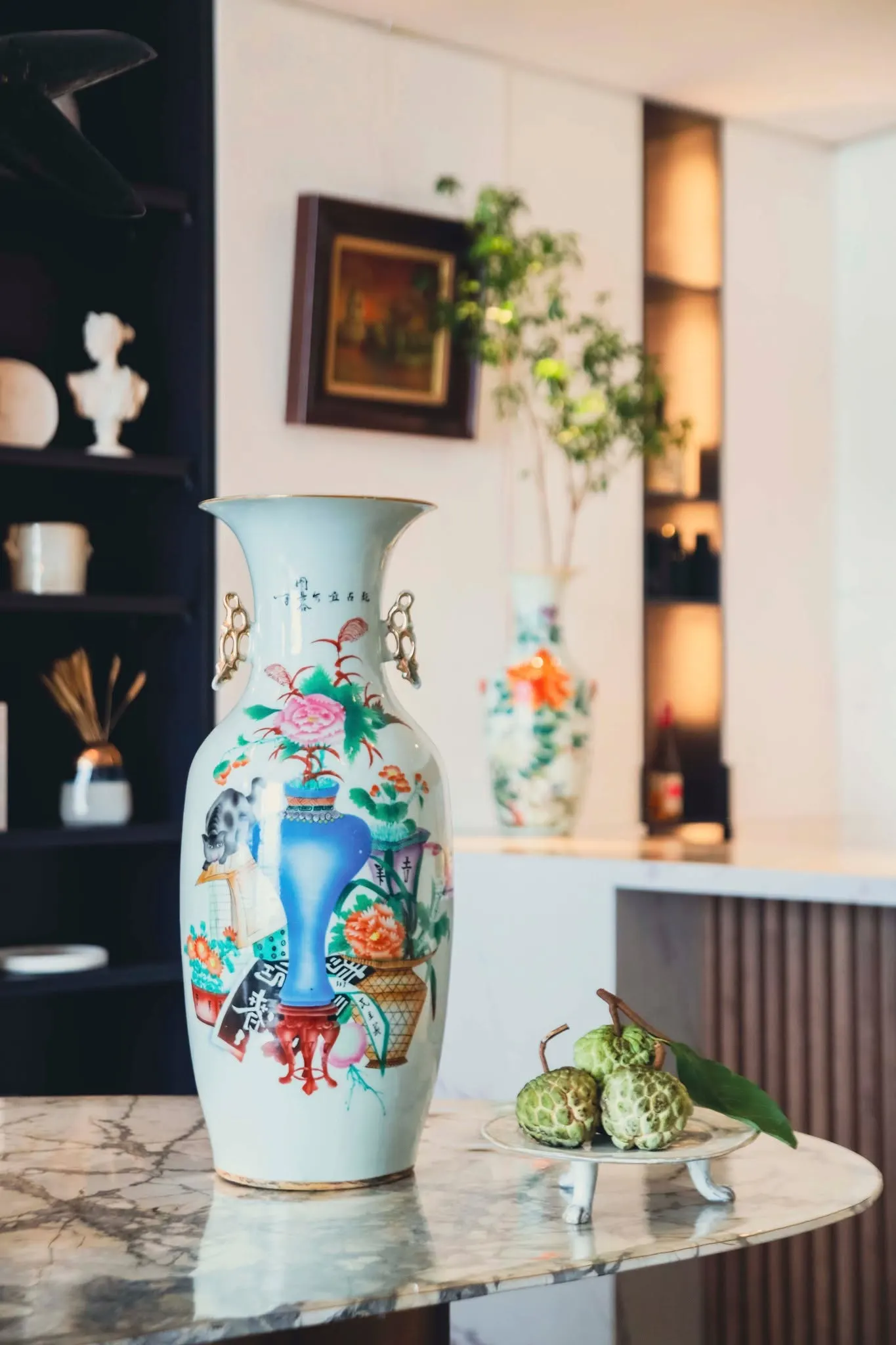 A tall painted porcelain vase and green custard apples on a marble table in front of dark shelves.