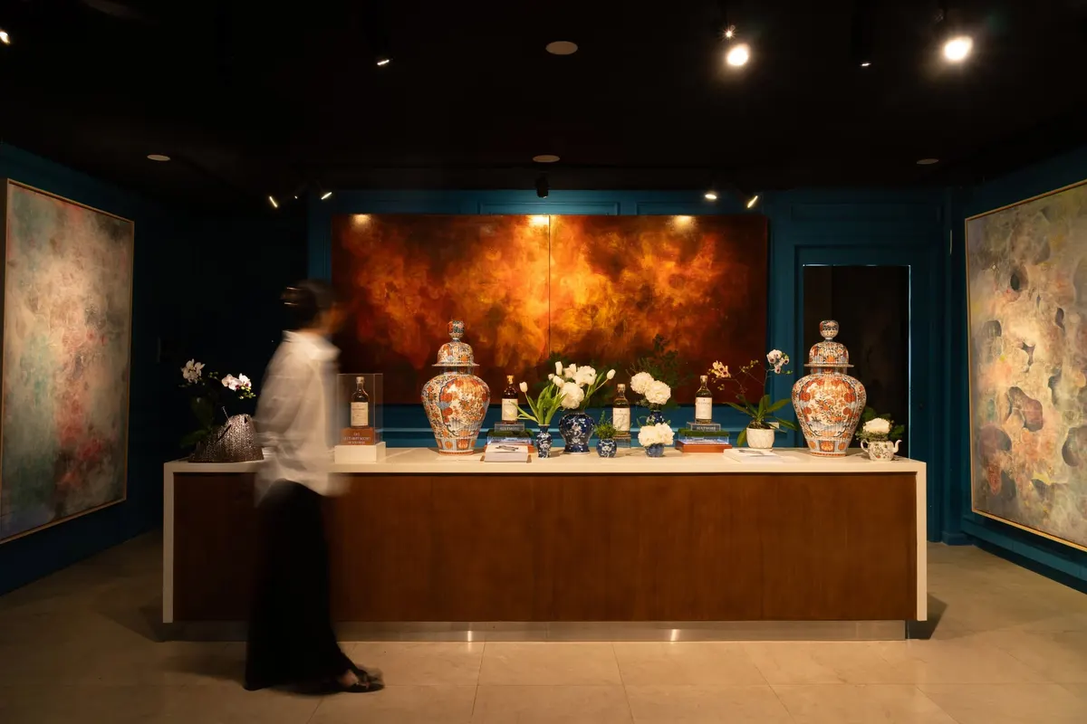 A dark gallery room at Indochine House: a glowing amber lacquer painting on a teal wall above a long counter with two painted lidded vases, white flowers and whisky bottles, a blurred figure in white walking past, large abstract paintings on both sides.