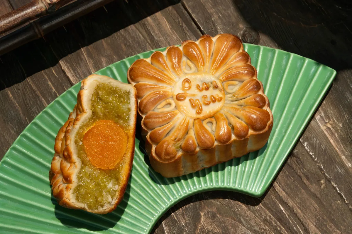 A baked Lam Yên mooncake with the brand name written on top, beside a half mooncake showing green cốm and a golden egg yolk, on a fan-shaped green plate on a dark wooden table.
