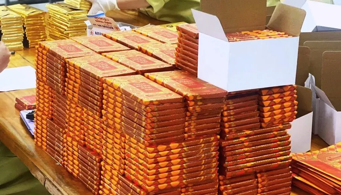 Tall stacks of orange-and-red chocolate bars piled up on a long wooden table in the workshop, a white box open at the top right.
