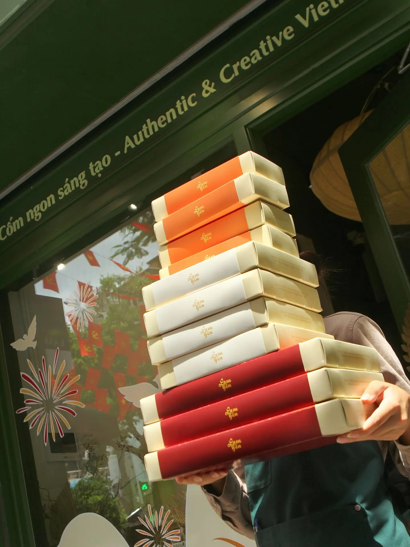 A hand holding a stack of red Lam Yên gift boxes with cream edges, in front of a shop window.